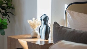 Ein Schlafzimmer mit einem Bett, einem Nachttisch mit Lampe, einer Vase mit Trockenblumen und einer Pflanze.