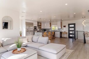 A modern living room with a sectional sofa, a dining area with wooden chairs, a kitchen with stainless steel appliances, and a staircase with a metal railing. - Modern 5BR Oasis - City Views, Gym & Gated Parking (San Diego)