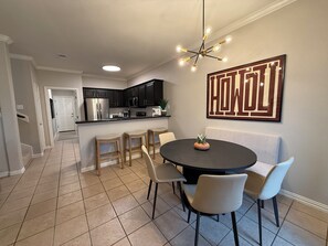 A dining area with a round table, four chairs, a chandelier, and a kitchen in the background.
