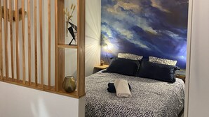 A bedroom with a bed, a nightstand, a wall-mounted shelf with decorative items, and a large wall mural depicting a cloudy sky. - Quiet studio at the foot of St Jacques hill,a stone's throw from the city center (Cavaillon)