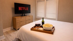 A bedroom with a TV displaying 'NETFLIX,' a wooden dresser, a tray with wine glasses, a bottle, and popcorn on a bed. - Modern Butterfly - Stylish 3 Bed 3 Bath with Hot Tub! Rumbling Bald Amenities! (Lake Lure)