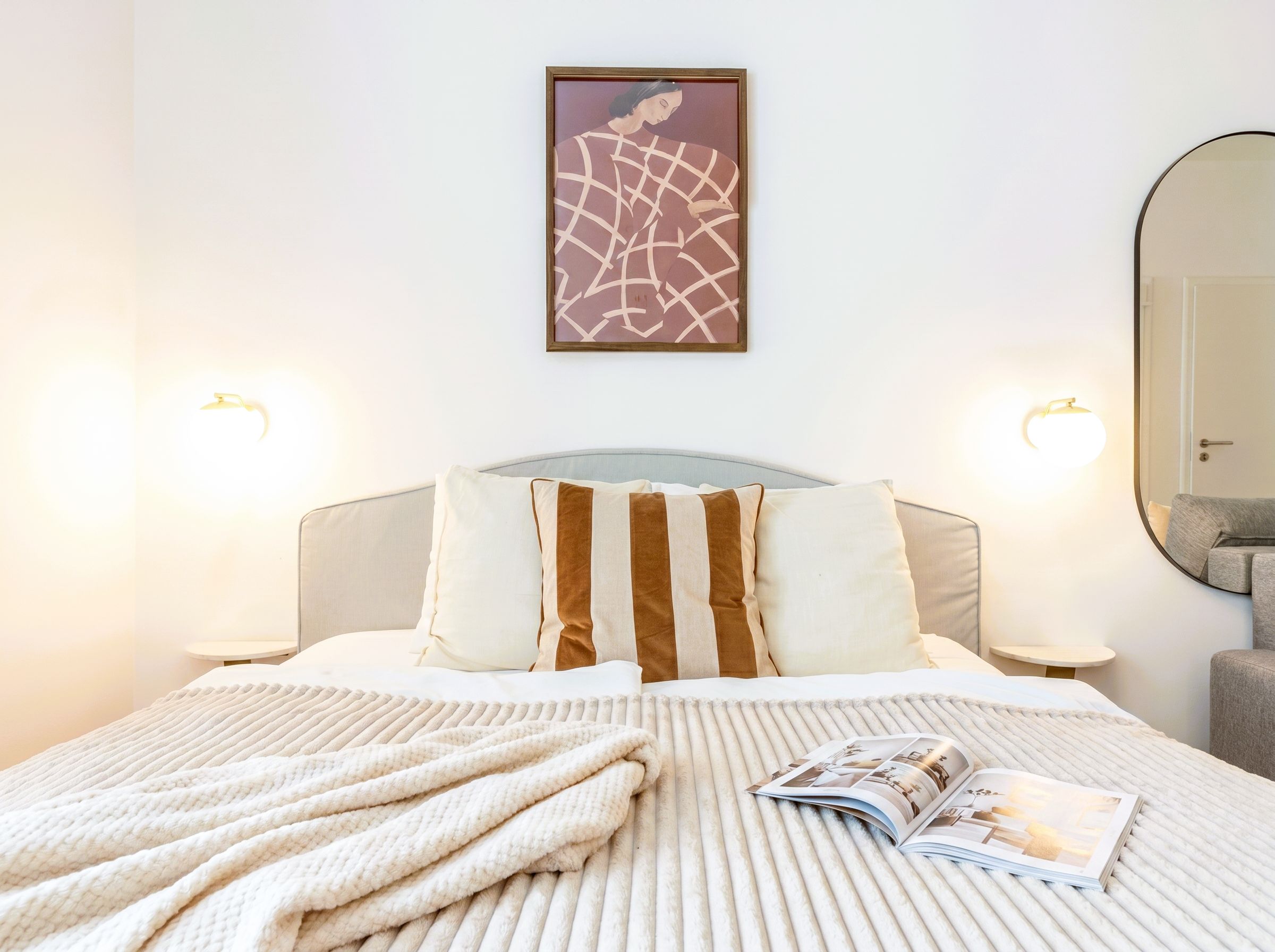 A hotel room with a bed, pillows, a blanket, a magazine, a framed artwork, and wall-mounted lights.
