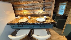 A rustic cabin interior with a wooden log wall, a wooden dining table with two chairs, a small kitchenette, and a window.