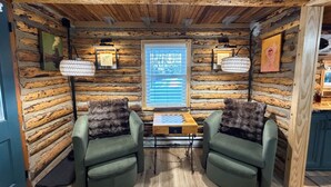 Interior de una cabaña de madera con dos sillones verdes, una mesa de madera con un tablero de ajedrez y una ventana con persianas.