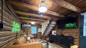 A log cabin interior with a leather sofa, a patterned rug, a fireplace, a staircase, and a neon sign.