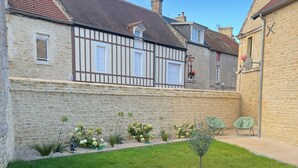 A courtyard with stone walls, a grassy area, and a building with a tiled roof and timber framing. - Maison proche mer (Reviers)