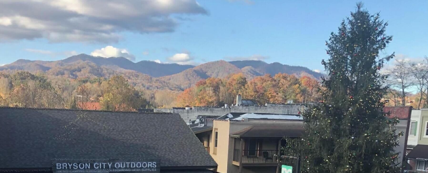 A mountain range with autumn foliage, a building with a sign for Bryson City Outdoors, and a Christmas tree.