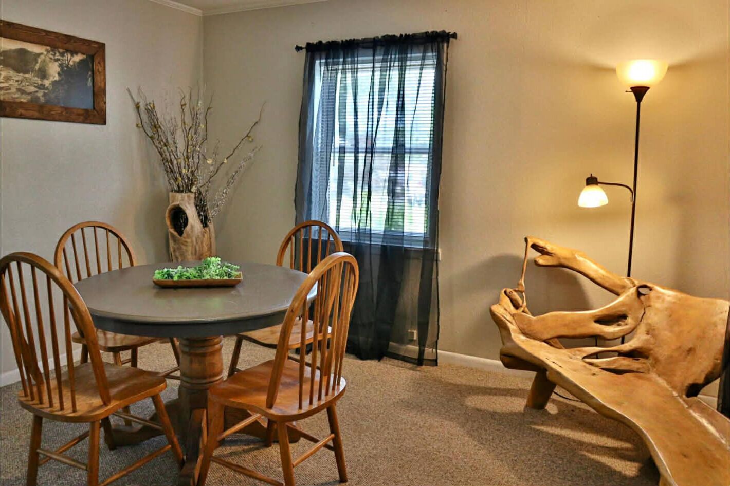 A dining area with a round table, wooden chairs, a vase with branches, a window with sheer curtains, and a wooden sculpture.