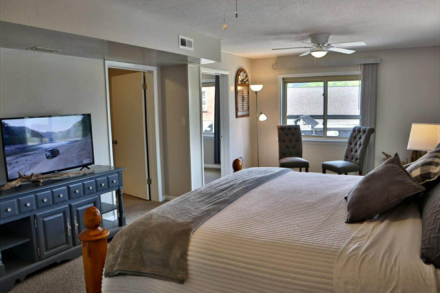 A hotel room with a bed, a television on a dresser, a ceiling fan, and a window with a view.