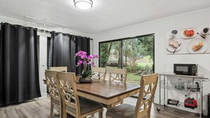 A dining area with a wooden table, chairs, a vase of flowers, a view through large windows, and a kitchen area with a microwave and storage cart. - Forest River Farmhouse Retreat (Fall Creek)