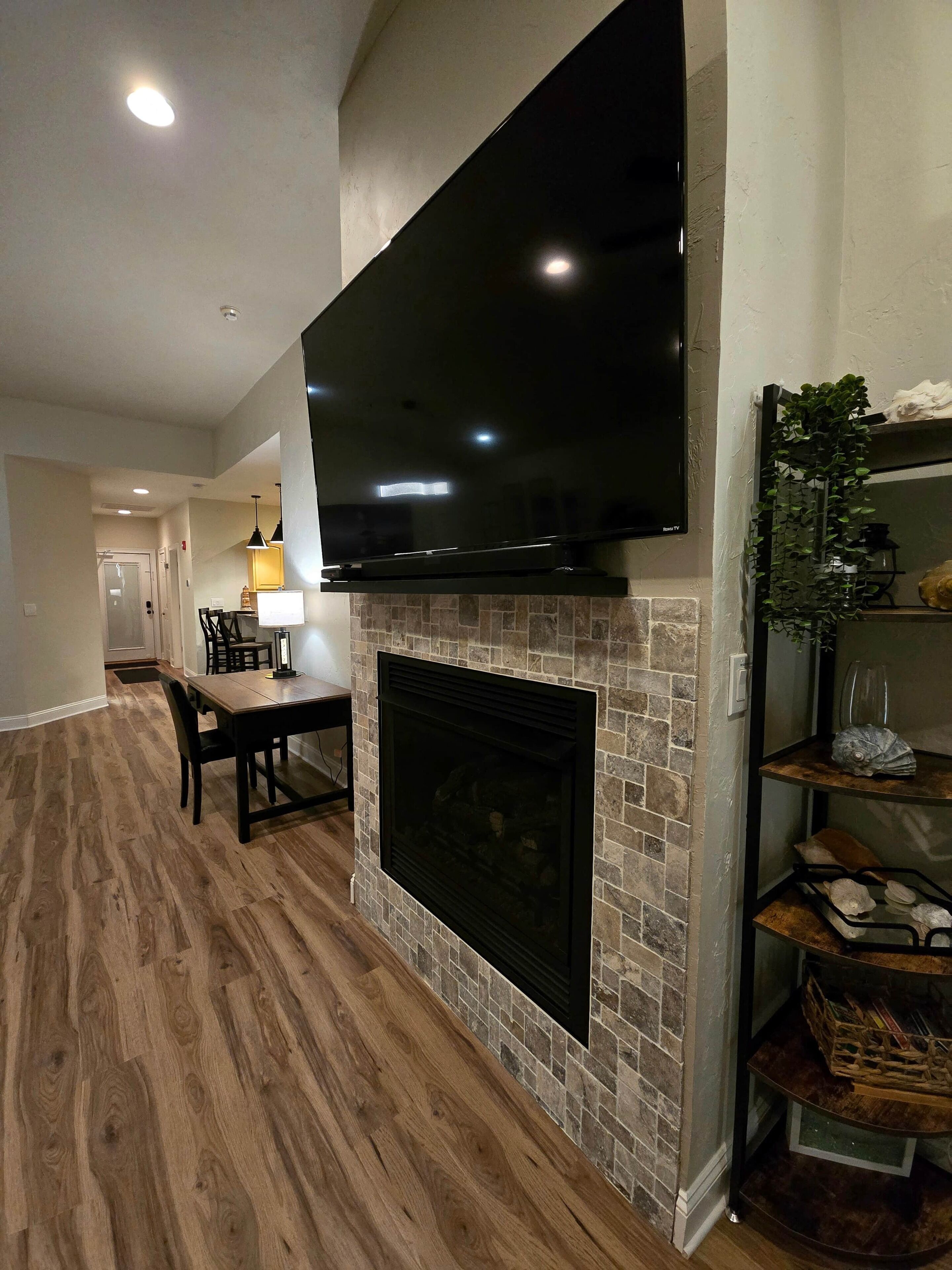 A modern living room with a flat-screen TV mounted above a fireplace, a dining area with a wooden table and chairs, and a bookshelf with decorative items.