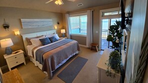 A bedroom with a bed, bedside lamps, a ceiling fan, a painting, a window, and a balcony.