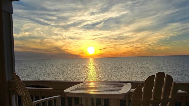 A wooden deck with a round table and chairs facing a sunset over the water.