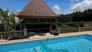 Piscina transparente com espreguiçadeira flutuante, um gazebo e balaustrada de pedra.