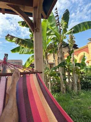 Eine Hängematte unter einer Holzkonstruktion, im Hintergrund Bananenbäume und ein Gebäude.