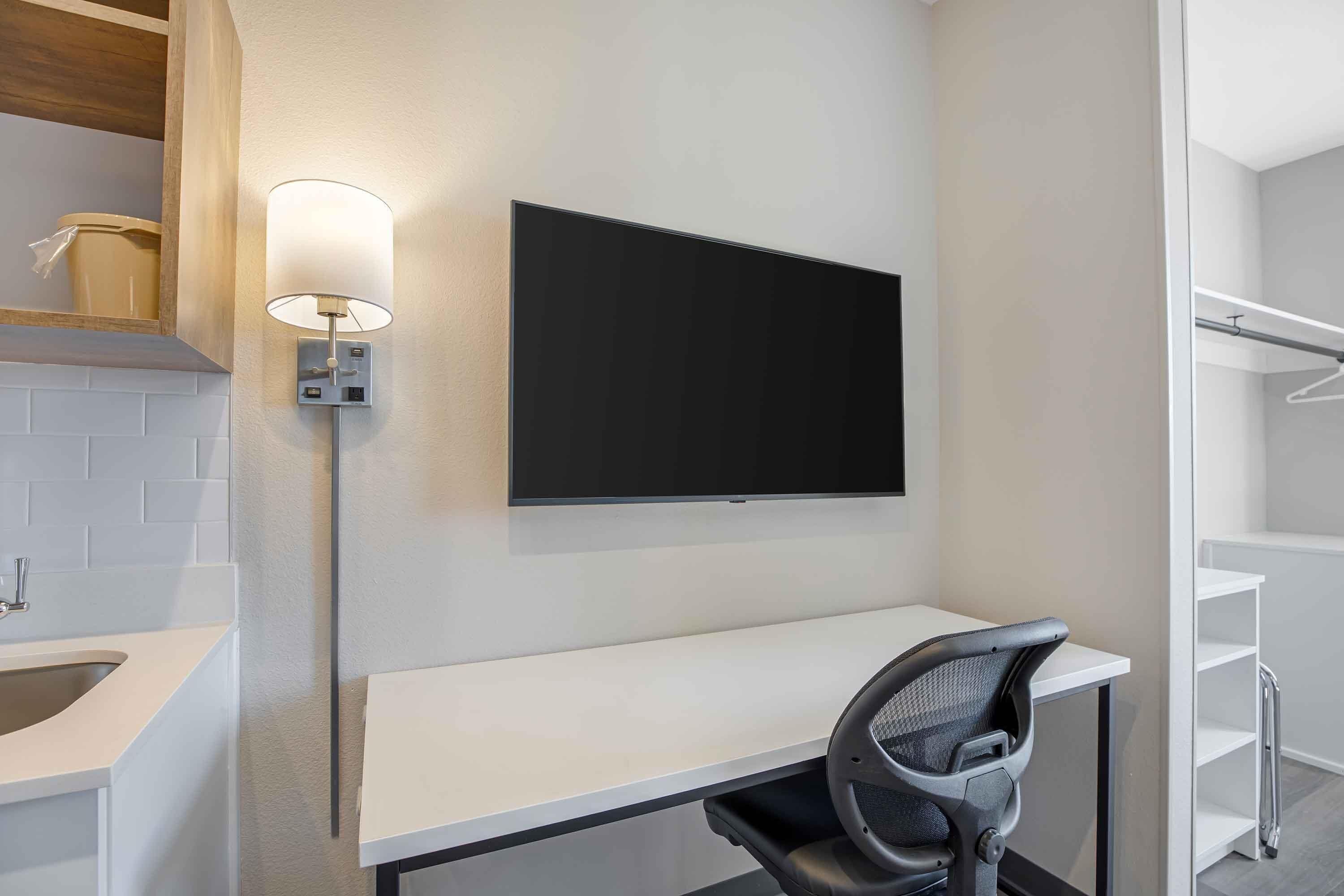 A modern room with a white desk, a black chair, a mounted flat-screen TV, and a wall-mounted light fixture.