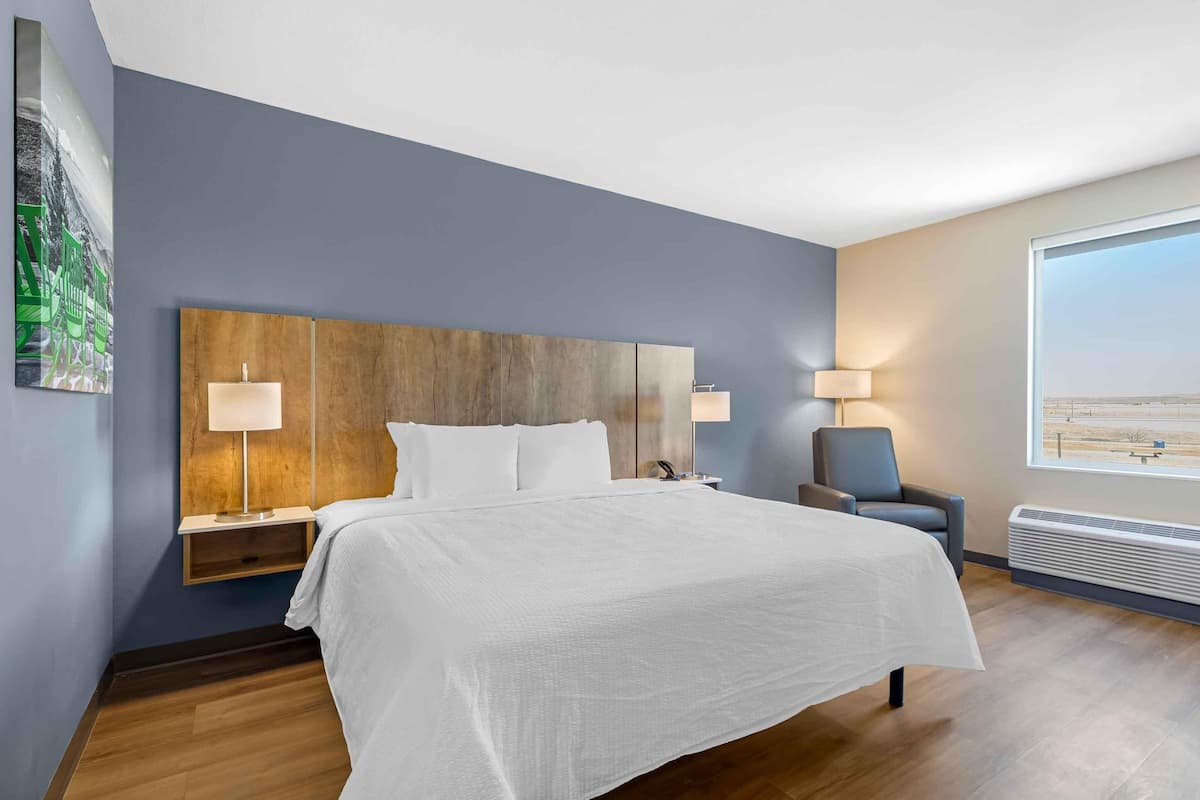 A hotel room with a large bed, bedside lamps, a chair, a nightstand, a wall-mounted heater, and a large window with a view.