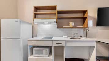 Eine kleine Küche mit weißen Geräten, einer Mikrowelle und einem Spülbecken. Über der Arbeitsplatte befinden sich Holzregale und ein Flachbildfernseher ist an der Wand montiert.