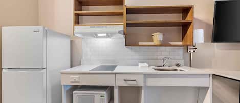 A compact kitchen with white appliances, a microwave, and a sink. There are wooden shelves above the counter and a flat-screen TV mounted on the wall.