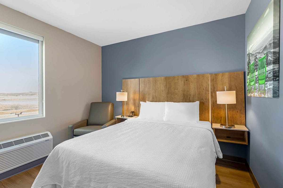 A hotel room with a large bed, two bedside tables with lamps, a chair, a window with a view, and a wall-mounted artwork.