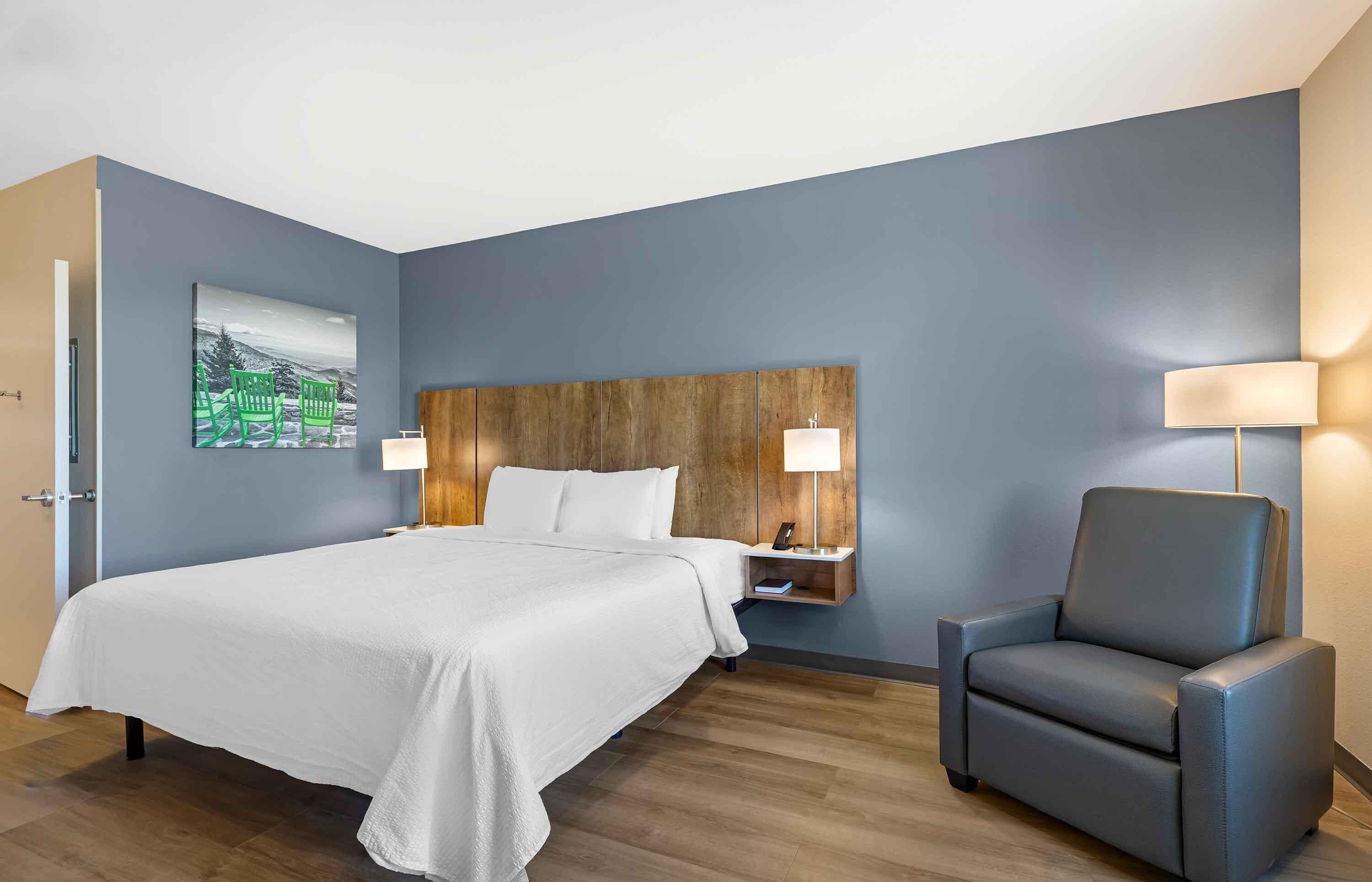 Habitación de hotel con cama, una silla, una mesita de noche con lámpara y estante en la pared.
