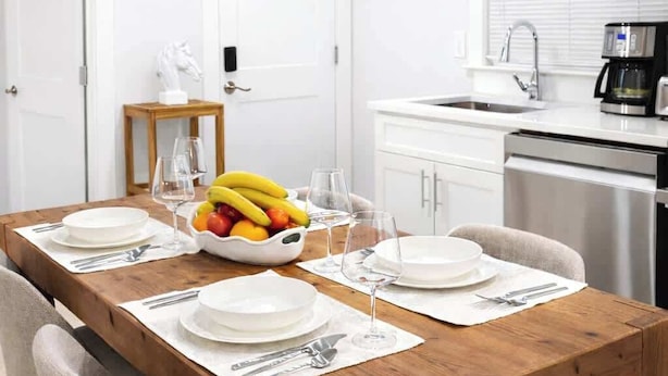 A dining area with a wooden table set for four, white chairs, a fruit bowl, and a kitchen in the background.