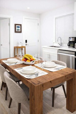 A dining area with a wooden table set for four, white chairs, a fruit bowl, and a kitchen in the background.