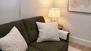 A living room with a green sofa, white cushions, a lamp, and a wall-mounted air conditioner.