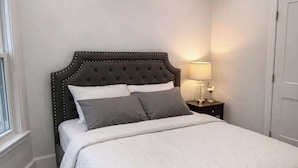 A bedroom with a bed, a bedside table with a lamp, an air conditioning unit, and a window.