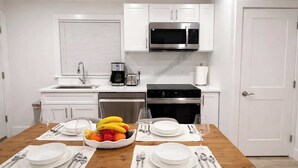 A modern kitchen with white cabinets, a microwave, a coffee maker, and a dining table set for two with plates, cutlery, and a fruit bowl.