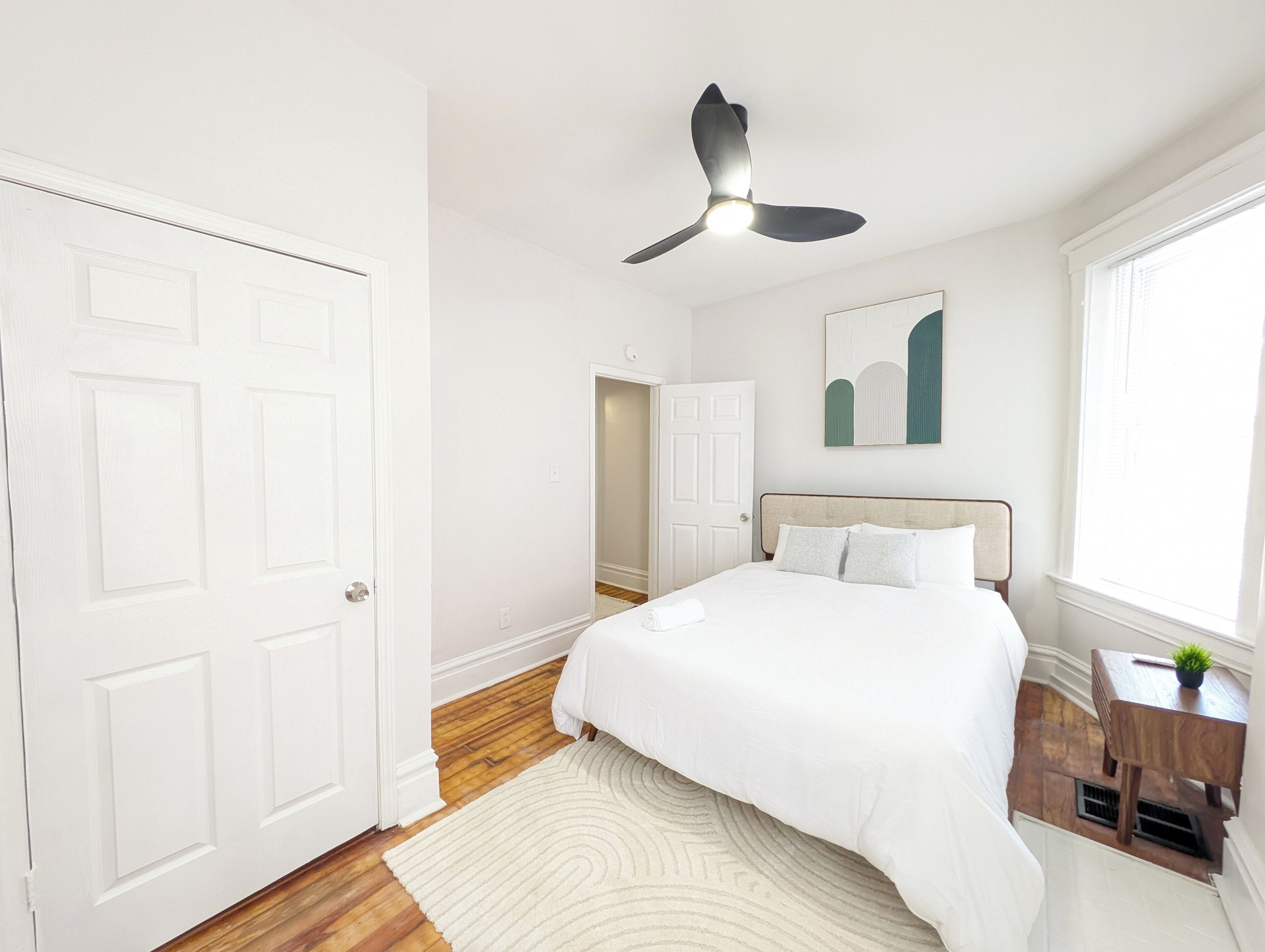 A bedroom with a bed, a ceiling fan, a window, a door, and a small table with a plant.
