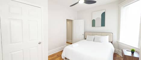 A bedroom with a bed, a ceiling fan, a window, a door, and a small table with a plant.