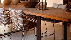 Un comedor con una mesa de madera, sillas de mimbre, un cuenco de piedra y portavelas.