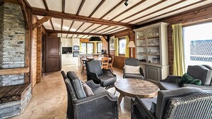 Un interior acogedor con chimenea de piedra, vigas de madera, muebles de mimbre, un comedor y una cocina al fondo.
