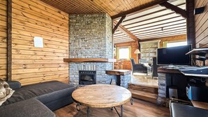 Un interior de cabaña acogedor con chimenea de piedra, paredes de madera, un sofá, una mesa redonda y un televisor.