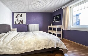 A bedroom with a bed, a radiator, a shelf, and a window with curtains.