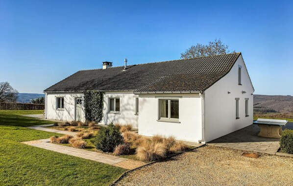 A white house with a tiled roof, a front garden with grass and shrubs, and a clear sky.