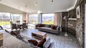 Ampla sala de estar com sofá, mesa de centro e mesa de jantar. Janelas grandes oferecem vista para o exterior.