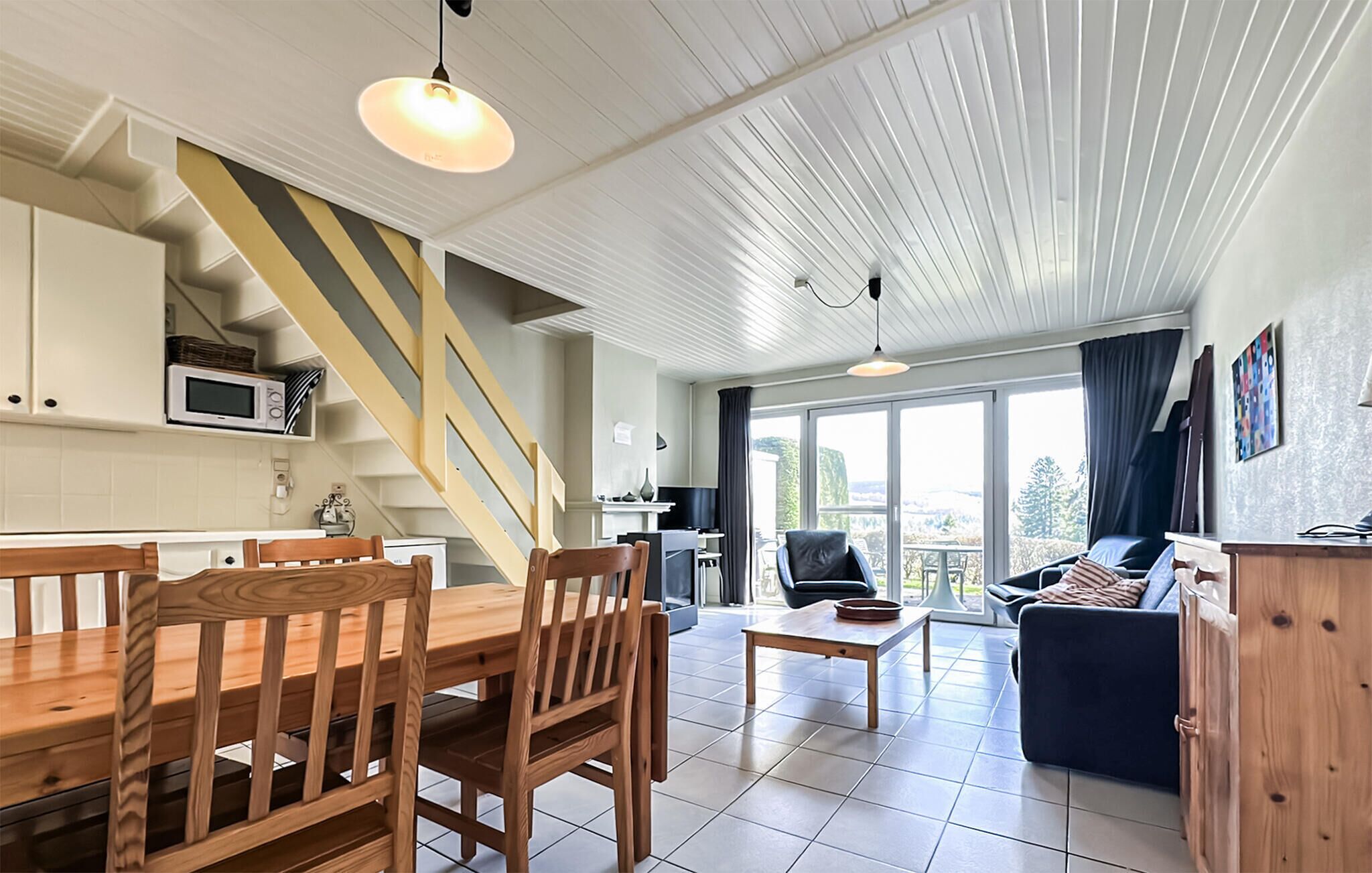 A modern interior with a dining area, kitchen, and living space, featuring a staircase, large windows, and a view of the outdoors.