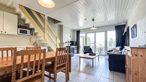 Un intérieur moderne comprenant un coin repas, une cuisine et un espace de vie, avec un escalier, de grandes fenêtres et une vue sur l’extérieur.