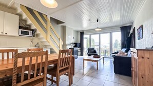 A modern kitchen with a wooden staircase, a dining area with wooden furniture, and a living room with a sofa and a view through large windows.