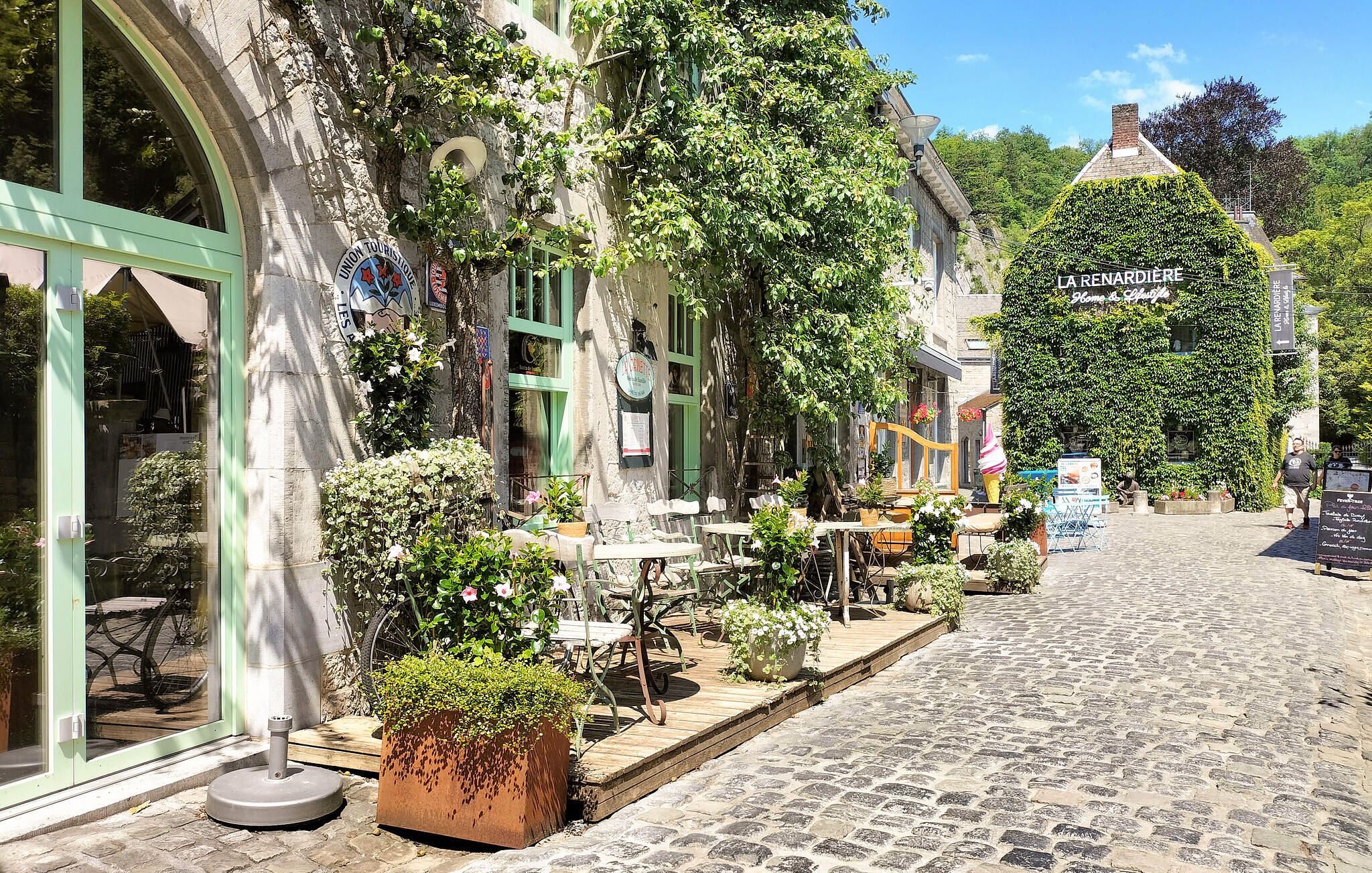 Rua de paralelepípedos com mesas ao ar livre, um edifício com a placa 'LA RENARDIE' e vegetação.