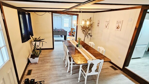A dining area with a long wooden table, white chairs, a potted plant, and a door leading to another room.