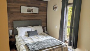 A bedroom with a wooden headboard, a bed with a plaid bedspread, a gray throw blanket, a nightstand with a lamp, a clock on the wall, and a window with curtains. - Cozy cottage located just over an hour to St. John's, NL! Pets welcome! (Placentia)