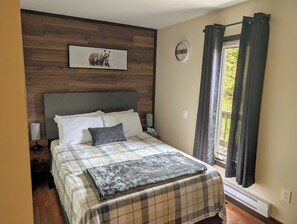 A bedroom with a wooden headboard, a bed with a plaid bedspread, a gray throw blanket, a nightstand with a lamp, a clock on the wall, and a window with curtains.