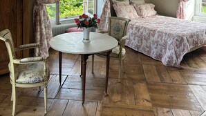 A room with a wooden floor, a round table with a vase of flowers, a bed with floral bedding, and a chair with a floral pattern. - Charming 17th-Century French Manor (Le Breuil-en-Bessin)
