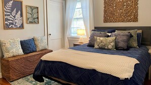 Quarto com cama, mesa de cabeceira, abajur, um objeto decorativo na parede e duas gravuras botânicas emolduradas.