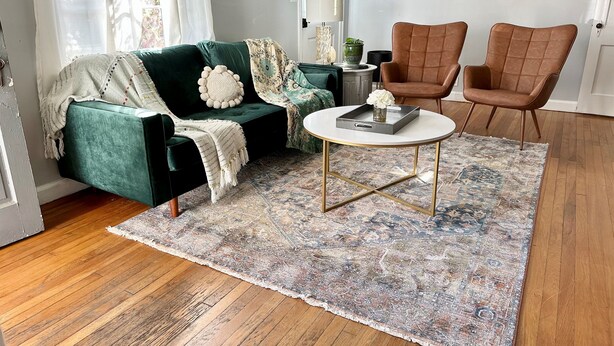 Uma sala de estar com um sofá verde, duas poltronas marrons, uma mesa de centro redonda e um tapete estampado.