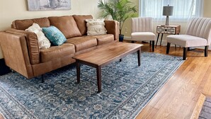 Ein Wohnzimmer mit braunem Ledersofa, einem Holztisch, einem blauen Teppich mit Muster, einer Topfpflanze und einem weißen Sessel.