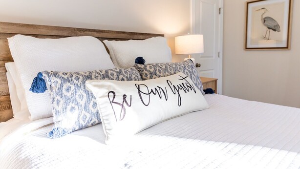 Un lit avec une literie blanche et des coussins décoratifs, une tête de lit en bois, une table de chevet avec une lampe et un tableau encadré au mur.